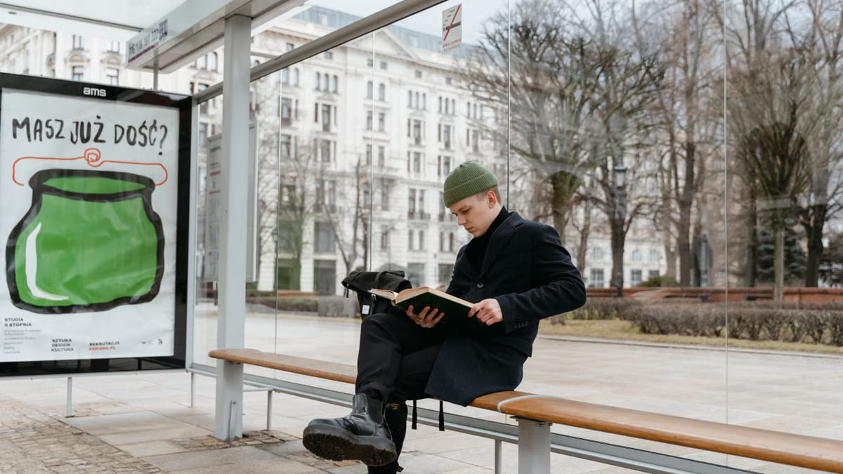 A man in a beanie and black coat reads at an outdoor bus stop with an advertisement.